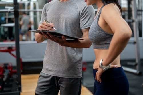 CloseUpFitnessTrainerCoachManWithClipboardTalkingTo - SomethingBeautiful!! Fitness trainer with clipboard discusses a workout plan with a woman in a gym setting.