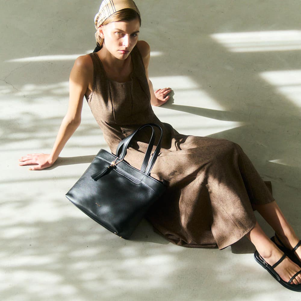 12_000000000765 - SomethingBeautiful!! Model in a brown sleeveless dress sits on a concrete floor with a black handbag resting beside her.