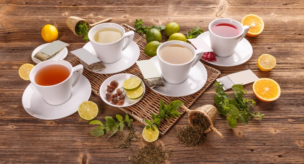 FourTeacupsWithDifferentTeasHerbsFruitsAndTeaBags - SomethingBeautiful!! Tea tasting setup: five white cups of tea on a wooden table with citrus slices, mint, and sugar nearby on a wicker mat.”