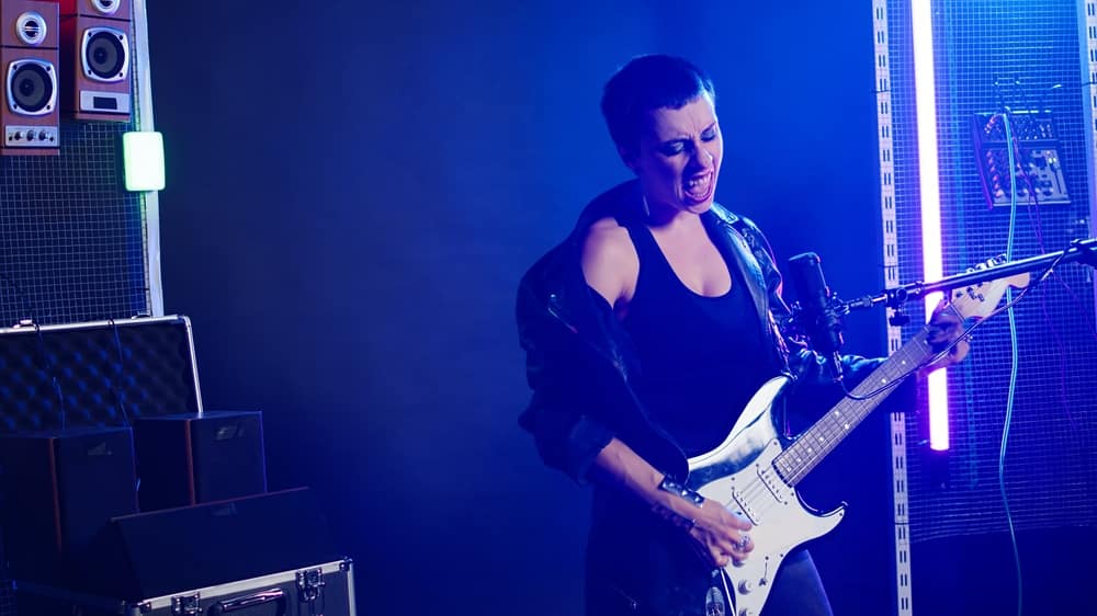 TalentedFemaleGuitaristDeliversAPowerfulMusicPerformanceInA - SomethingBeautiful!! Female guitarist passionately performing in a neon-lit studio, playing a white electric guitar while singing into a microphone with blue/purple lighting behind her.