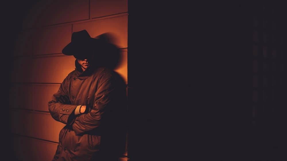 MysteriousPortraitInTheNoirStyleOfAMaleDetective - SomethingBeautiful!! Person wearing a wide-brim hat and coat leaning against an orange-lit wall in a dim night scene.