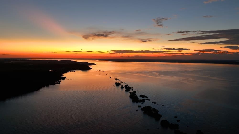 SunsetAerialViewAtAmazonRiverOfAmazonRainforestAt - SomethingBeautiful!! Sunset over a calm river with a diagonal line of rocks stretching toward the horizon.