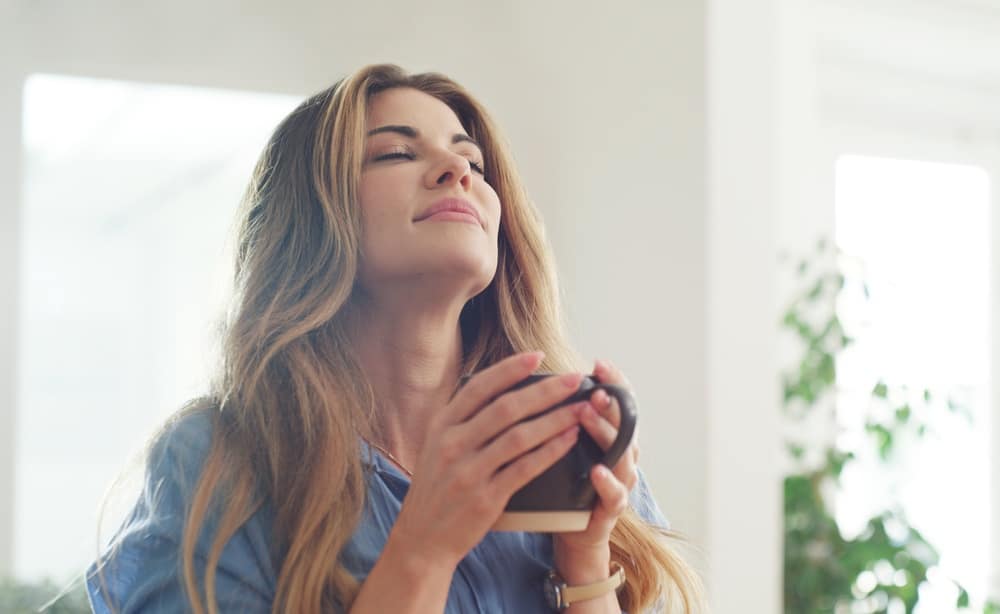 WomanSmellAndScentOfCoffeeAtHouseToStart - SomethingBeautiful!! Woman with long blonde hair and a blue shirt holds a mug close, eyes closed, enjoying a warm drink in a bright, airy room.