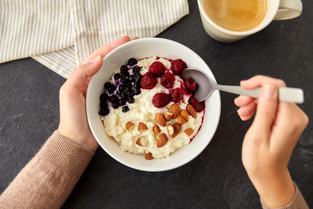 FoodAndBreakfastConcept-HandsOfWomanWithSpoon - SomethingBeautiful!! Person holding a bowl of yogurt with blueberries, raspberries, and almonds, spoon ready over a dark tabletop with a coffee cup nearby