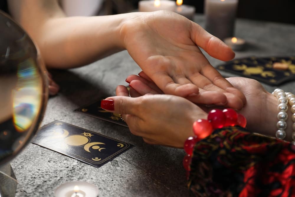 FortuneTellerReadingLinesOnWomansPalmAtBlackTable - SomethingBeautiful!! Close-up of two hands during a palm-reading session, with tarot cards on the table and candles burning in the background.