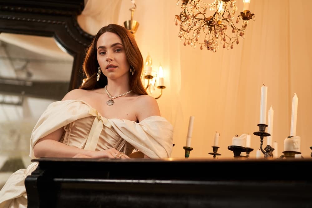 DrapedInAnElegantVintageDressAYoungWomanRadiates - SomethingBeautiful!! Elegant woman in an off-shoulder cream gown sits at a black piano, adorned with pearls and a chandelier-lit, candlelit room in warm light.