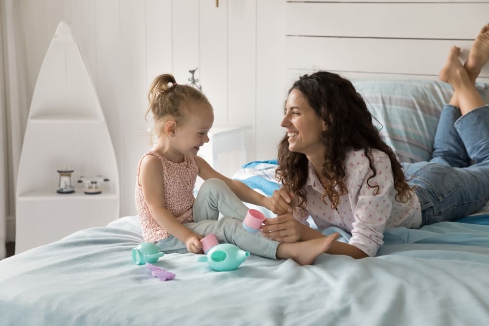 RolePlayingGameInChildDevelopmentSmilingYoungMotherSpend - SomethingBeautiful!! Mother and young daughter play tea party on a bed, sharing smiles and small pastel cups and teapot.