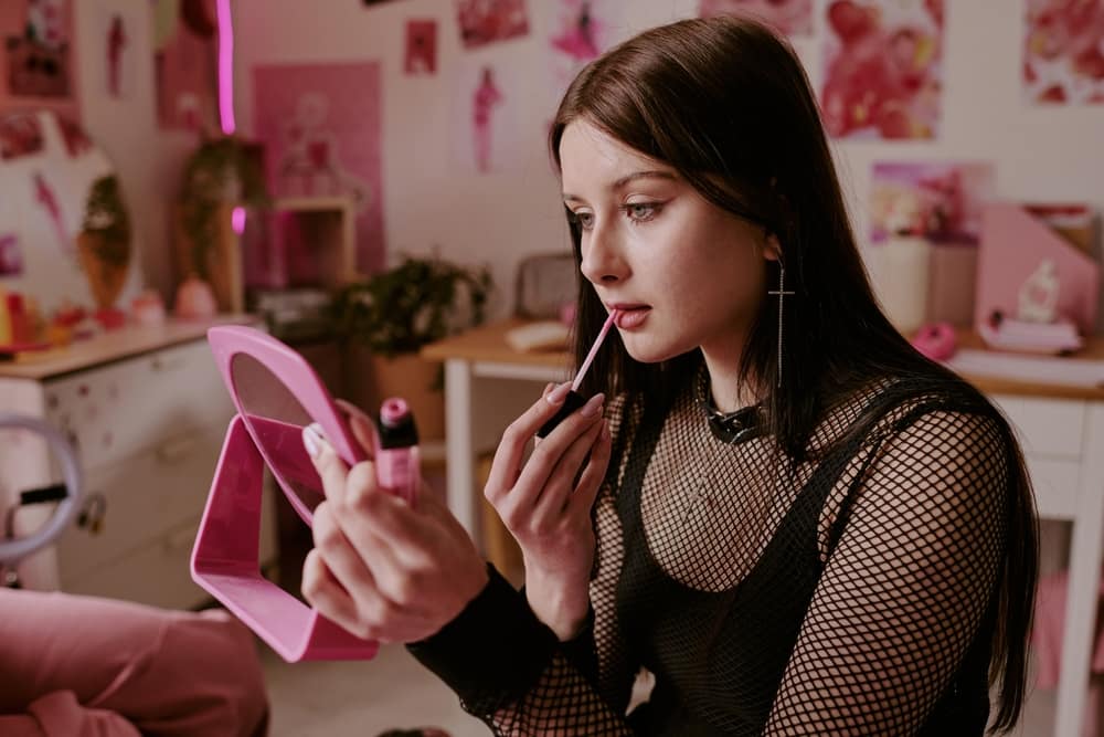 MediumCloseUpOfAdorableGothTeenInTotalBlack - SomethingBeautiful!! Young woman in a pink, decor-filled room applies pink lip gloss while looking into a pink handheld mirror.