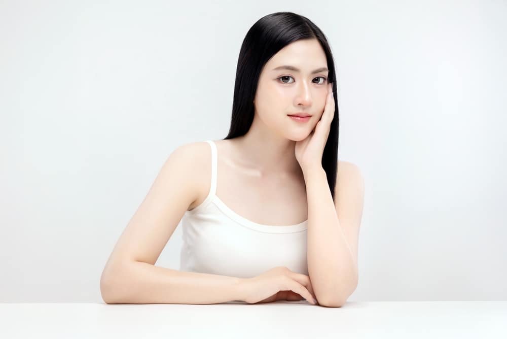 Young woman with long black hair resting her cheek on her hand at a white table in a bright studio environment, wearing a white tank top, calm expression.