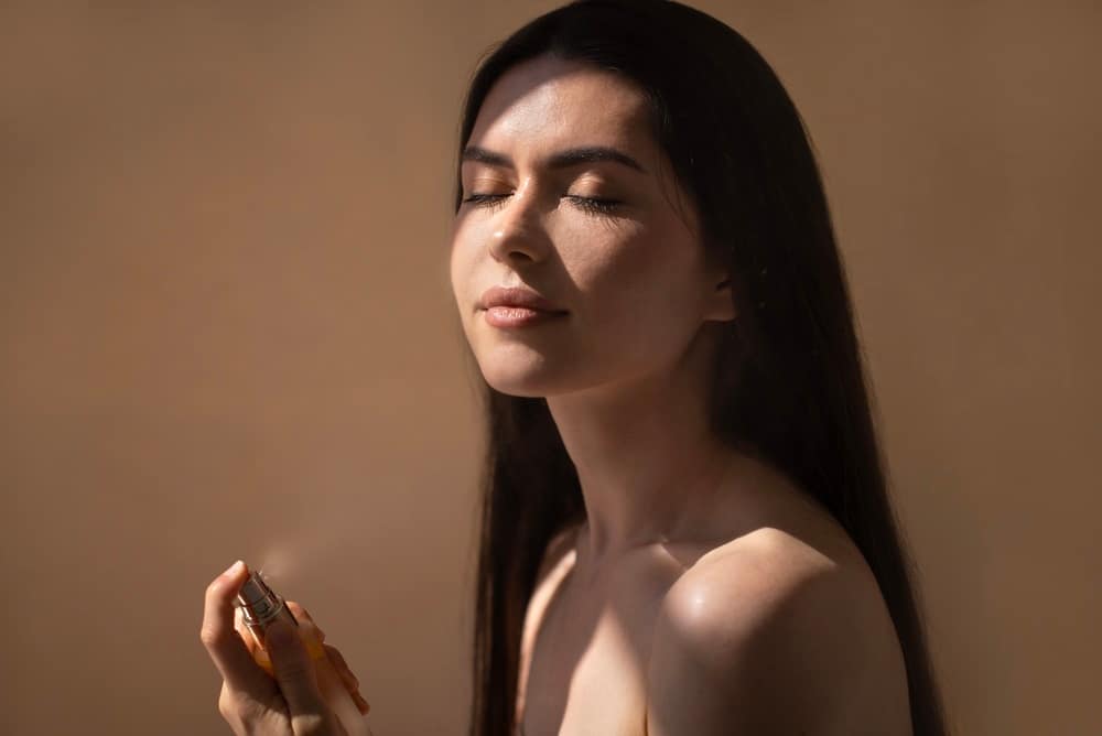 FemaleApplyingPerfumeWithLongDarkHairInSoftLight - SomethingBeautiful!! Woman with eyes closed in warm light, holding a perfume bottle near her shoulder.