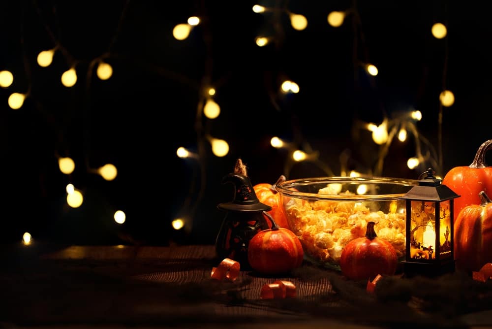AFestiveHalloweenPartySnackTableFeaturingAGlassBowl - SomethingBeautiful!! Autumnal centerpiece with small pumpkins, a crystal bowl of popcorn, a lit lantern, and warm string lights in the dark.