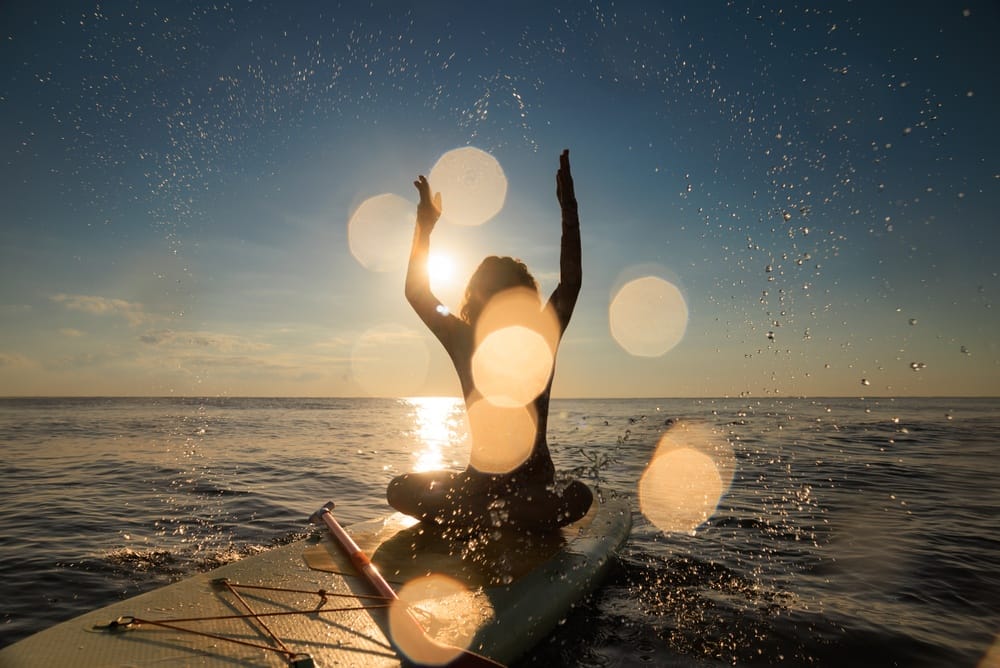 HappyWomanWalkingOnStand-upPaddleboardByTheSeaFemale - SomethingBeautiful!! Person on a paddleboard at sunset with arms raised and sunlit water droplets sparkling around them