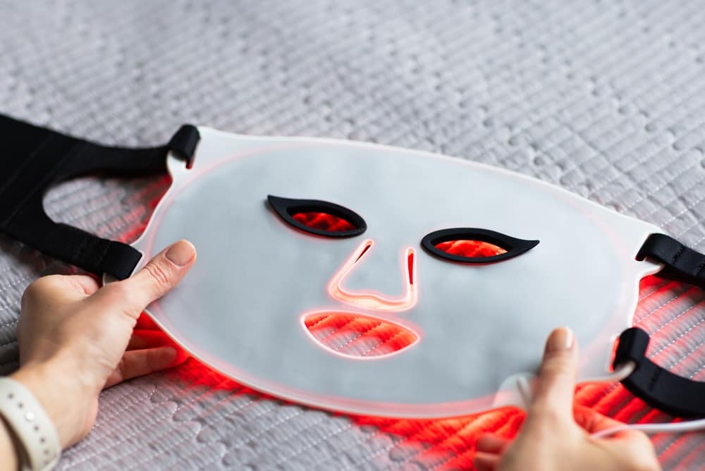 PersonHoldingLedFacialMaskAtHome - SomethingBeautiful!! Hands hold a white LED red-light facial mask on a textured surface, ready for therapy use.