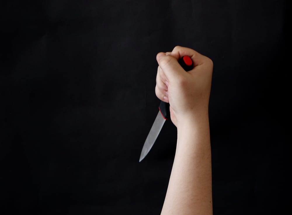 ManHandHoldingASharpAndCuttingKnifeOnBlack - SomethingBeautiful!! A hand gripping a knife with a red handle, blade pointing downward, against a dark background.