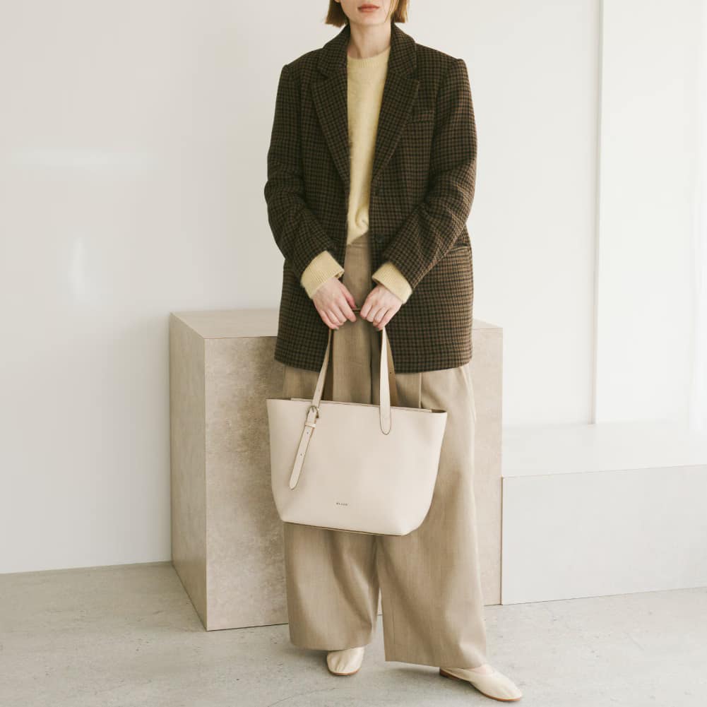 13_000000001358 - SomethingBeautiful!! Person standing in a neutral room wearing a brown check blazer, beige sweater, wide-leg beige pants, and holding a light beige tote bag.