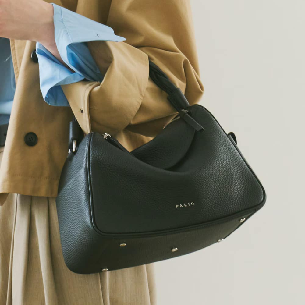 8_000000001353 - SomethingBeautiful!! Woman in a tan coat holding a black leather handbag with a blue shirt cuff visible, Palio bag with metal studs at the base