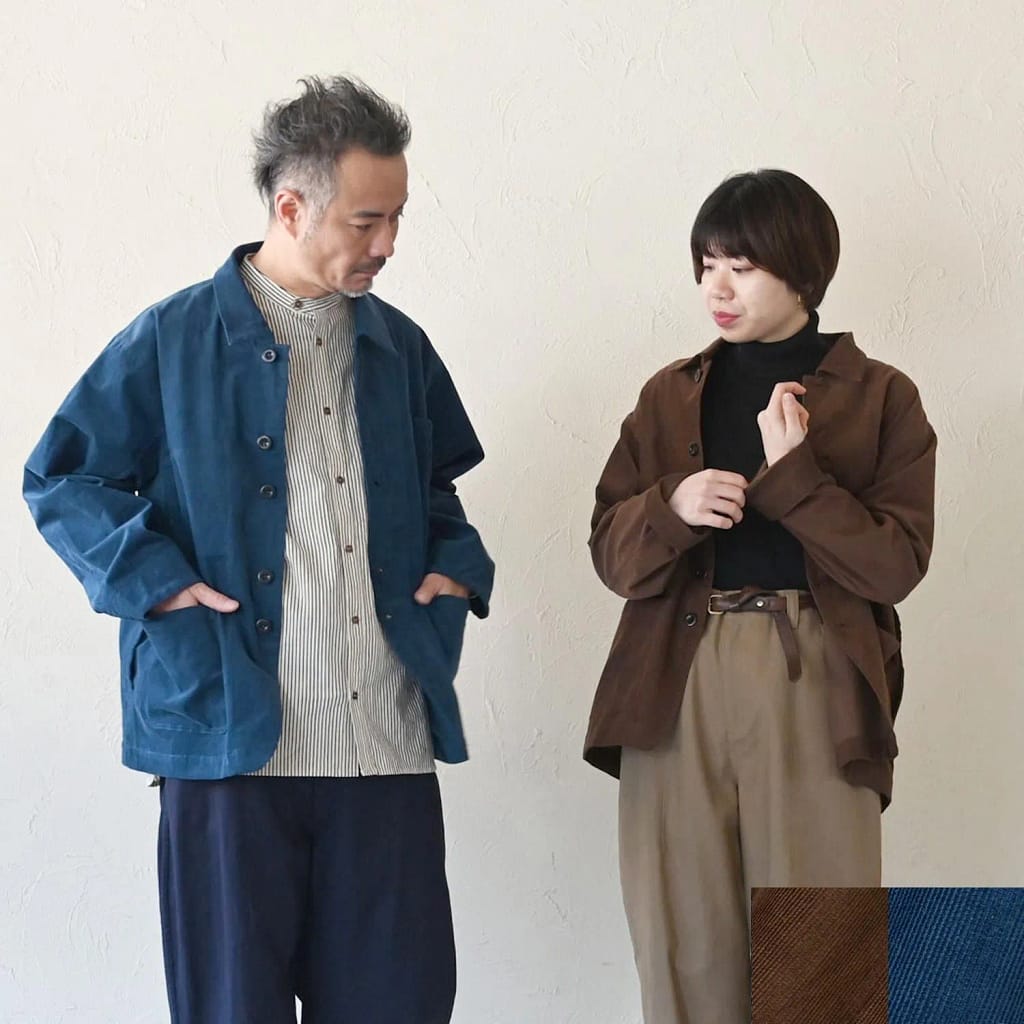 187379262 - SomethingBeautiful!! Two adults pose in a minimalist room, wearing layered outfits: a man in a blue jacket and striped shirt beside a woman in a brown overshirt and beige trousers against a white wall.