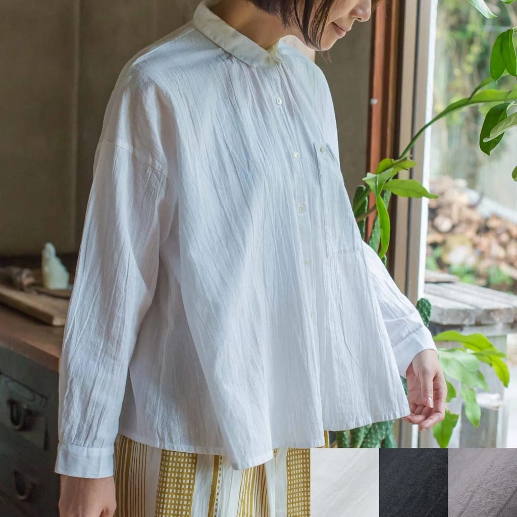 192560633 - SomethingBeautiful!! Person wearing a loose white button-up shirt standing near a window with green plants nearby.
