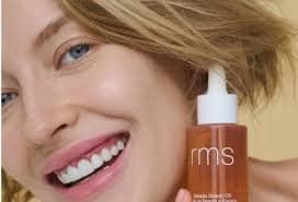 images 8 - SomethingBeautiful!! Close-up of a smiling woman with a pump bottle of RMS Beauty skincare held near her cheek.