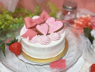 aba1dd9afd994bc383f5259806be7bb4-330x250 - SomethingBeautiful!! Small white frosted cake with pink heart cookies and swirls on a glass plate, surrounded by flowers and a strawberry.