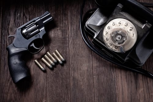 NoirFictionScene-HandgunWithCartridgesAndRotaryPhone - SomethingBeautiful!! Black revolver with six bullets laid out next to a vintage rotary dial telephone on a dark wooden surface.