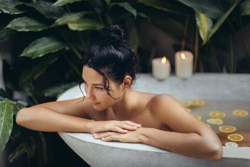ASereneWomanEnjoysATranquilBathInAnOutdoor - SomethingBeautiful!! Woman relaxing in an outdoor bathtub surrounded by lush plants, with candles lit nearby and lemon slices floating in the water