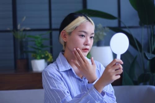 YoungAsianWomenLookingAtMirrorAndConcernAboutFacial - SomethingBeautiful!! Woman with blonde hair in a blue striped shirt holds a round hand mirror and examines her face, as if applying makeup, in a room with plants in the background.