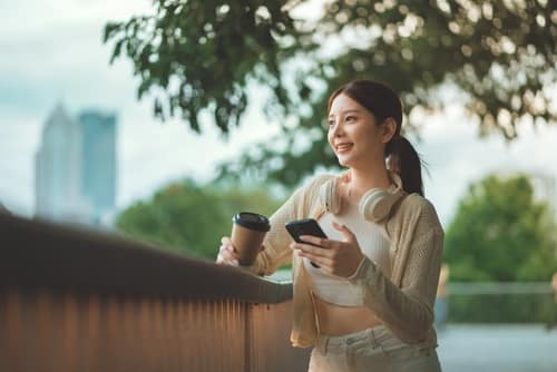 YoungAsianWomanEnjoyingARelaxingOutdoorMomentWithSmartphone - SomethingBeautiful!! Young woman outdoors, looking at her smartphone while holding a coffee cup and wearing headphones near a railing.