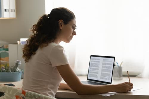 ConfidentFemaleInterpreterTranslateDocumentFromForeignLanguageToEnglish - SomethingBeautiful!! Woman with curly hair and glasses sits at a desk, typing on a laptop and taking notes on paper.
