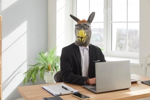 BusinessmanInDonkeyHeadMaskUsingLaptopComputerInOffice - SomethingBeautiful!! Person wearing a donkey mask and suit sits at a wooden desk with a laptop in a bright office space.