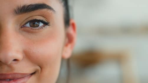 HalfFacePortraitIndianWomanSmilingLookingAtCameraIndoors - SomethingBeautiful!! Close-up of a young woman's face, showing her left eye, eyebrow, and a subtle smile.