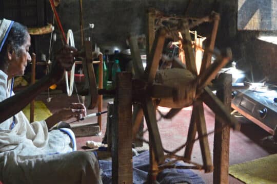 0519212-540x360 - SomethingBeautiful!! Elderly woman seated at a wooden loom, weaving fabric in a dim workshop.