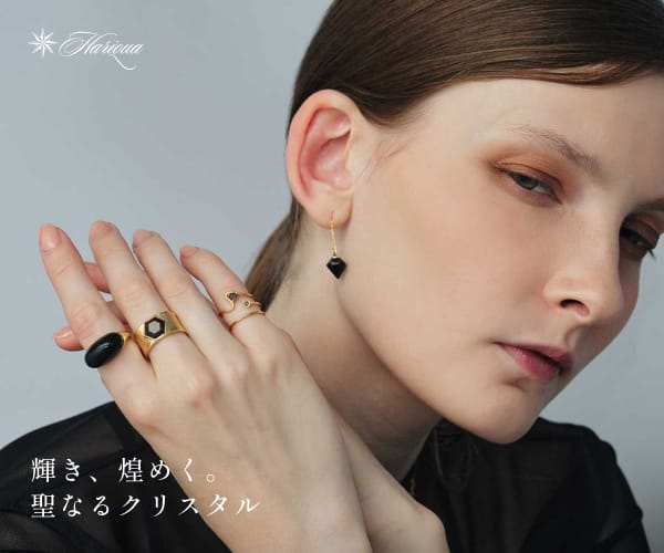 202411141014171810 - SomethingBeautiful!! Close-up of a woman showcasing gold jewelry—rings on her fingers and a drop earring—against a pale blue backdrop, with the brand slogan in Japanese in the corner.