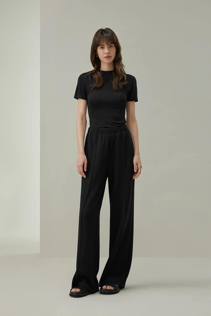 1 1 - SomethingBeautiful!! Full-body portrait of a woman in a black short-sleeve top and wide-leg pants standing against a neutral beige backdrop.