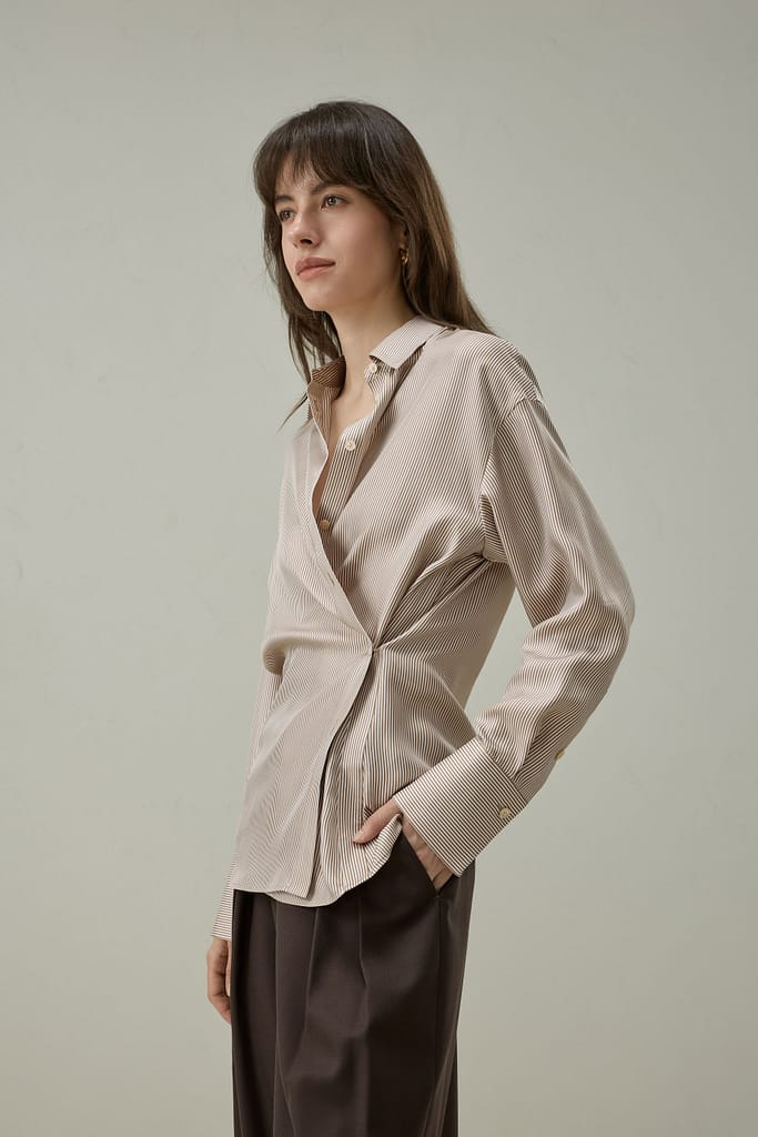 1 2 - SomethingBeautiful!! Woman in a beige striped button-up shirt and brown wide-leg pants, standing against a neutral wall with a thoughtful gaze to the left.