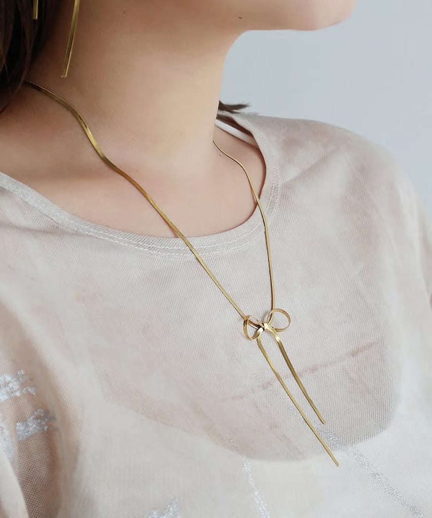 000000002459-02 - SomethingBeautiful!! Gold necklace with a bow pendant resting on a light beige top, close-up view